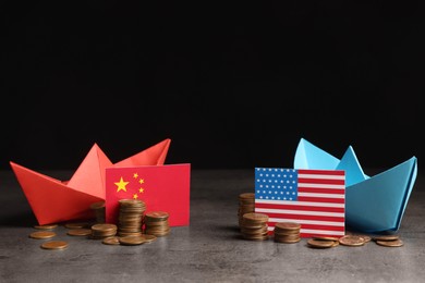 Photo of American and Chinese flags, coins with paper boats on grey table against black background . Trade war concept