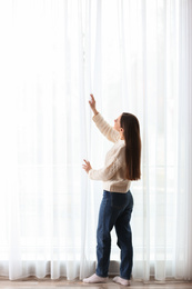 Young woman opening window curtains at home