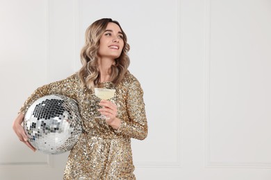 Photo of Beautiful woman in golden dress with glass of drink and disco ball posing near white wall indoors, space for text