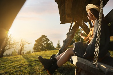 Photo of Woman with cozy plaid enjoying sunlight on outdoor swing in mountains