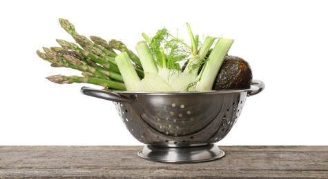 Metal colander with asparagus, fennel and avocados on wooden table against white background