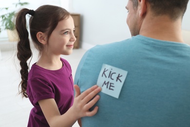 Cute little girl sticking note with words Kick Me to father's back at home