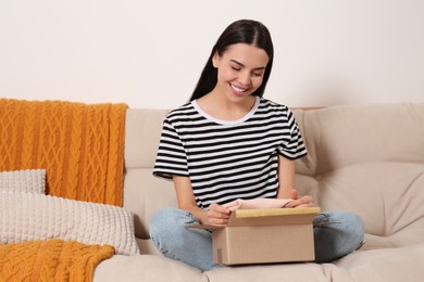 Happy young woman opening parcel on sofa at home. Internet shopping
