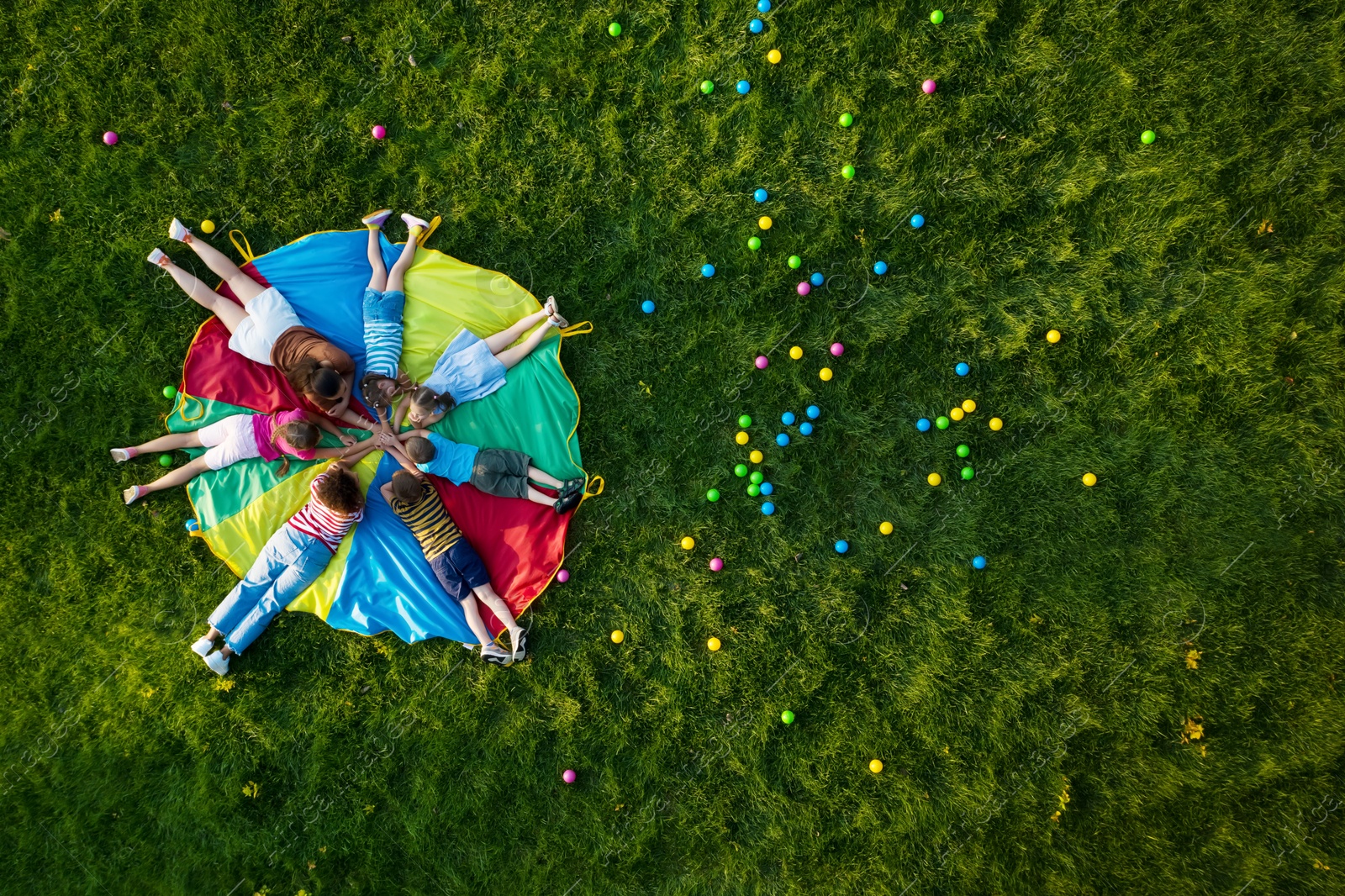 Image of Group of children with teachers holding hands together on rainbow playground parachute in park, top view. Summer camp activity