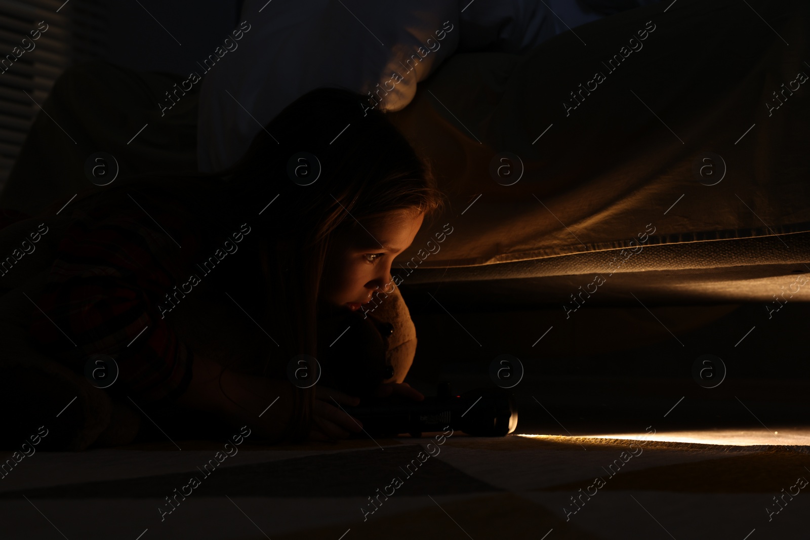 Photo of Little girl with flashlight looking for monster under bed at night