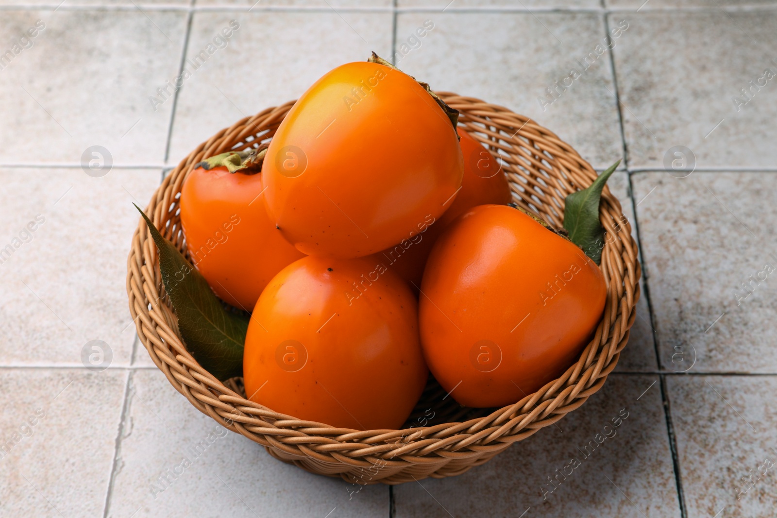 Photo of Delicious ripe juicy persimmons in wicker basket on tiled surface