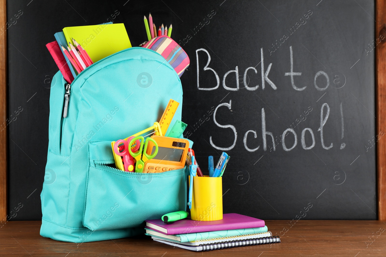 Photo of Bright backpack with school stationery on brown wooden table near black chalkboard. Back to School