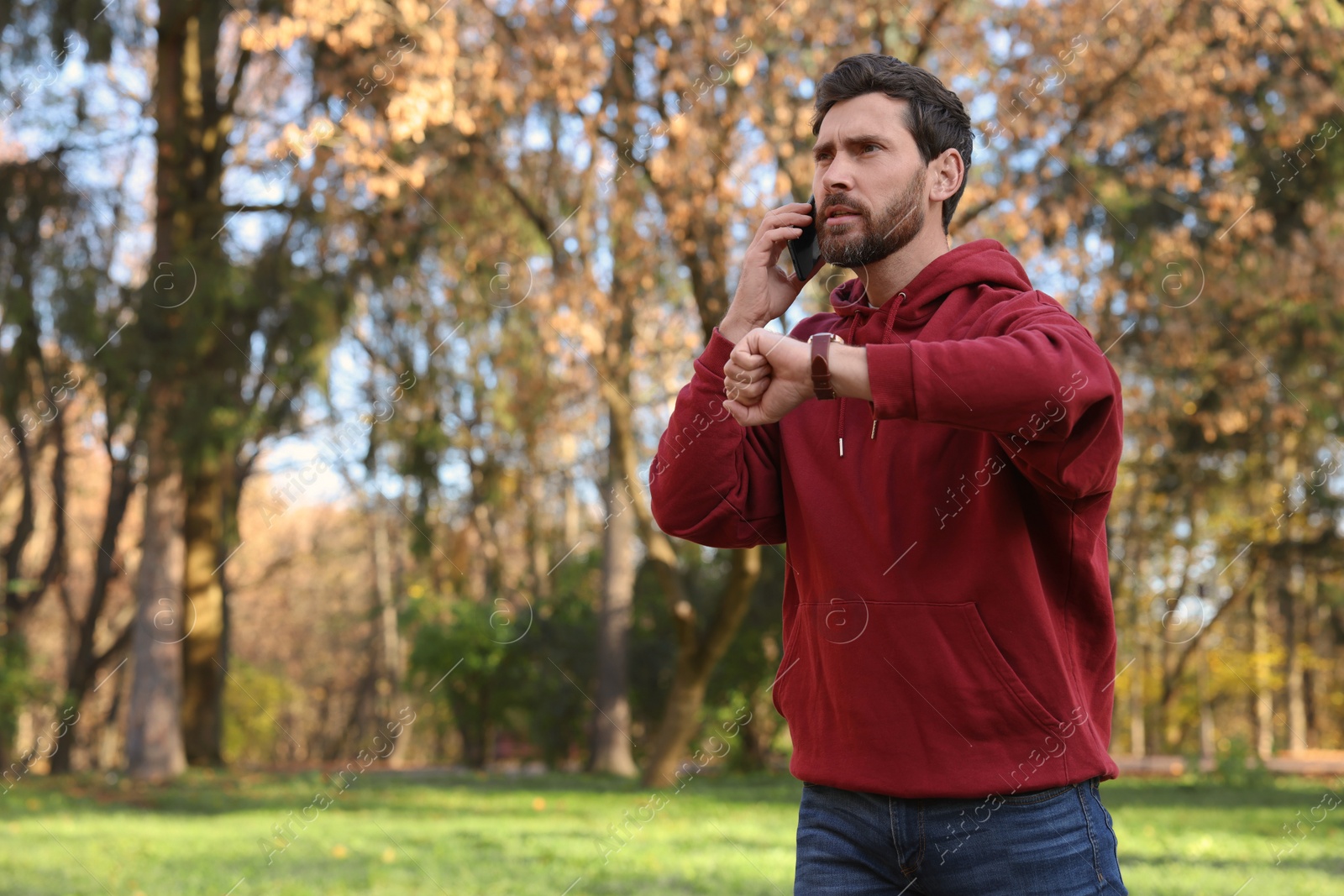 Photo of Emotional man talking by smartphone and checking time on watch in park, space for text. Being late concept