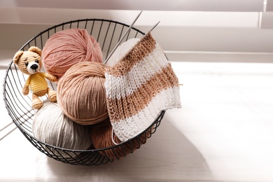 Yarn balls, knitting needles and cute handmade toy bear in metal basket on window sill indoors, space for text. Creative hobby