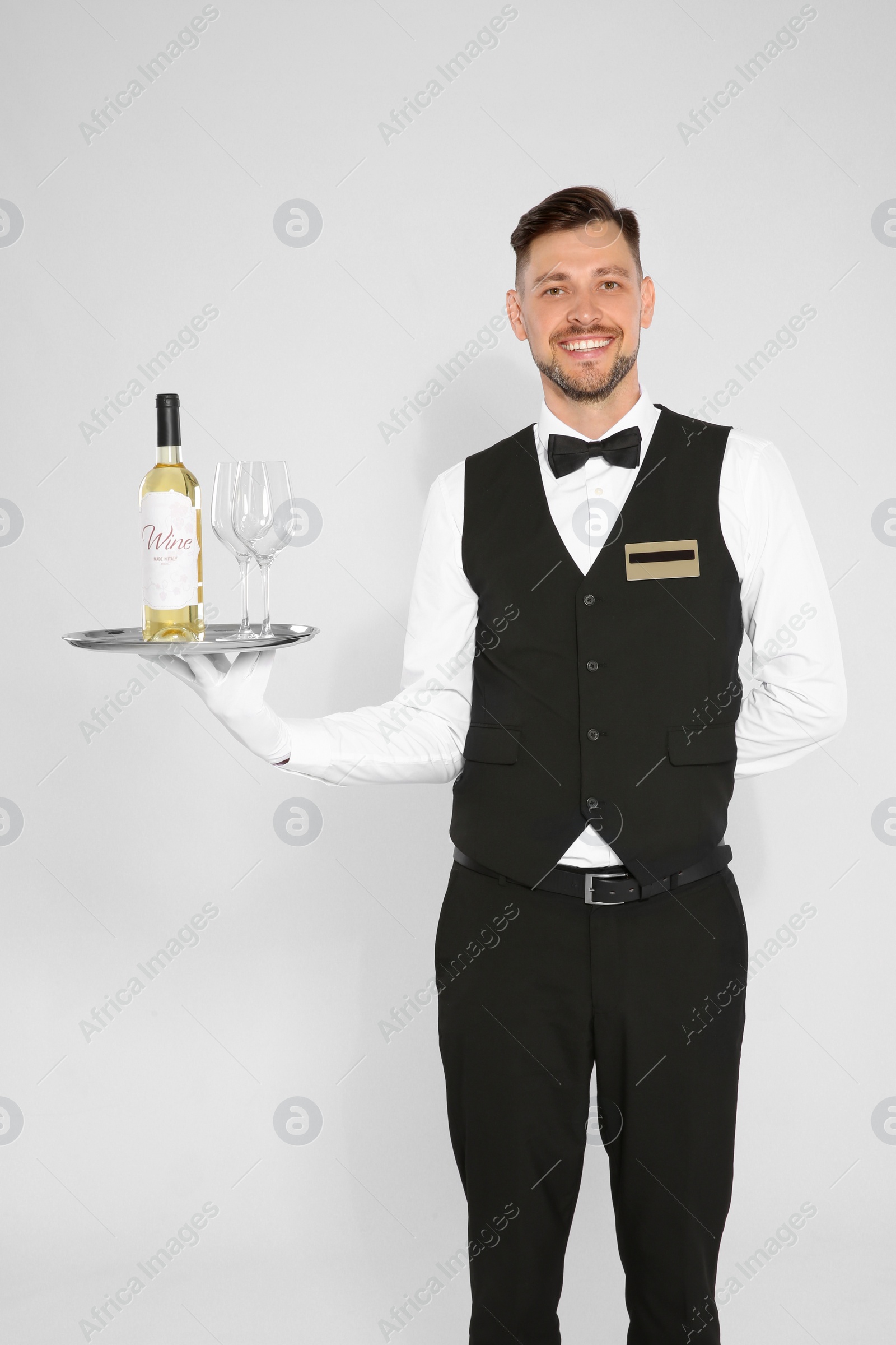 Photo of Handsome waiter holding tray with glasses and bottle of wine on light background