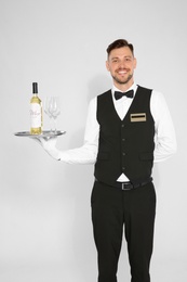 Photo of Handsome waiter holding tray with glasses and bottle of wine on light background