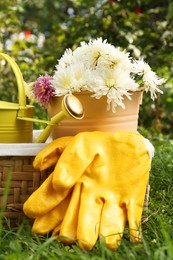 Wicker basket with gardening gloves, flowers and watering can on grass outdoors