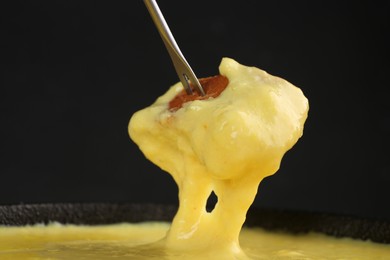 Dipping piece of bread into fondue pot with tasty melted cheese against dark gray background, closeup