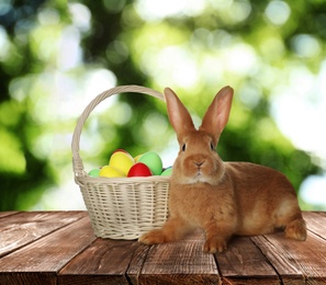 Adorable bunny and wicker basket with Easter eggs on wooden surface outdoors 