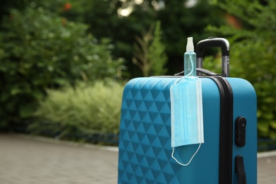 Photo of Stylish blue suitcase with protective mask and antiseptic spray outdoors, space for text. Travelling during coronavirus pandemic