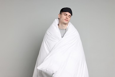 Man in pyjama and sleep mask wrapped in blanket on grey background