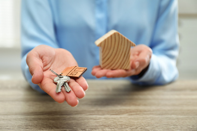 Real estate agent holding house figure and keys at wooden table, closeup