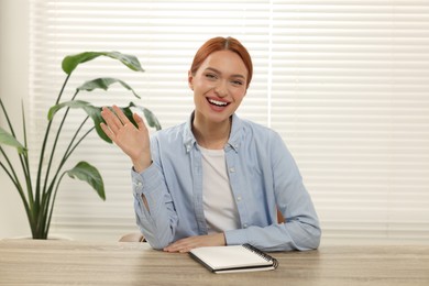 Young woman waving hello during video chat at wooden table indoors, view from web camera