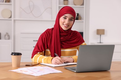 Muslim woman writing notes near laptop at wooden table at home