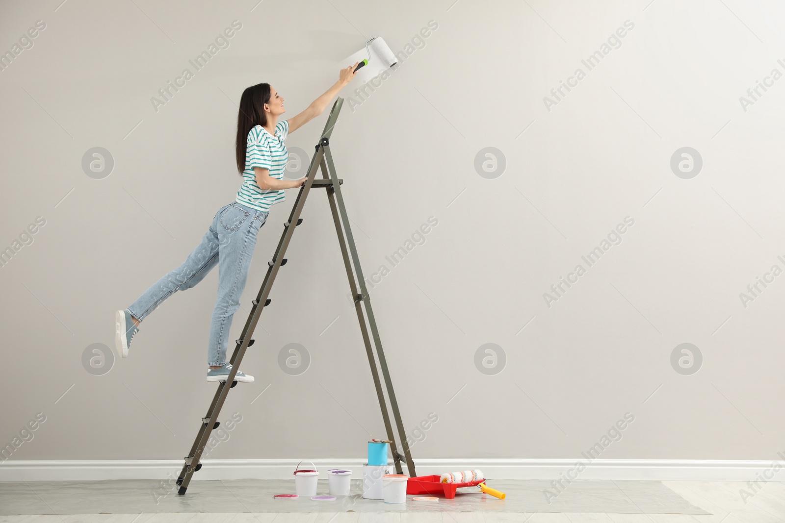 Photo of Young woman painting wall with roller on ladder indoors, space for text