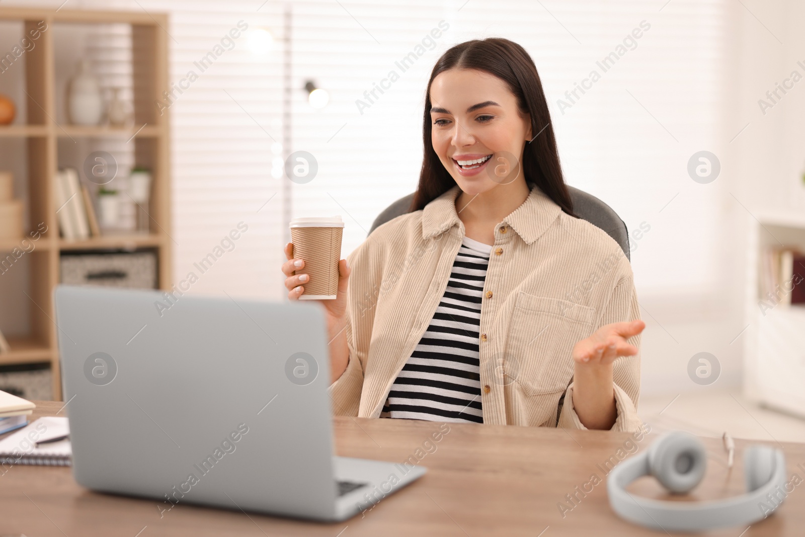 Photo of Young woman with cup of coffee using video chat during webinar at table in room