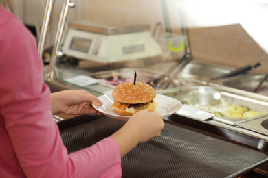 Photo of Little girl with plastic tray and burger near serving line in canteen, closeup. School food