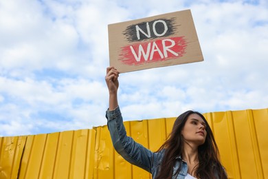 Photo of Young woman holding poster No War near yellow fence under cloudy sky. Space for text