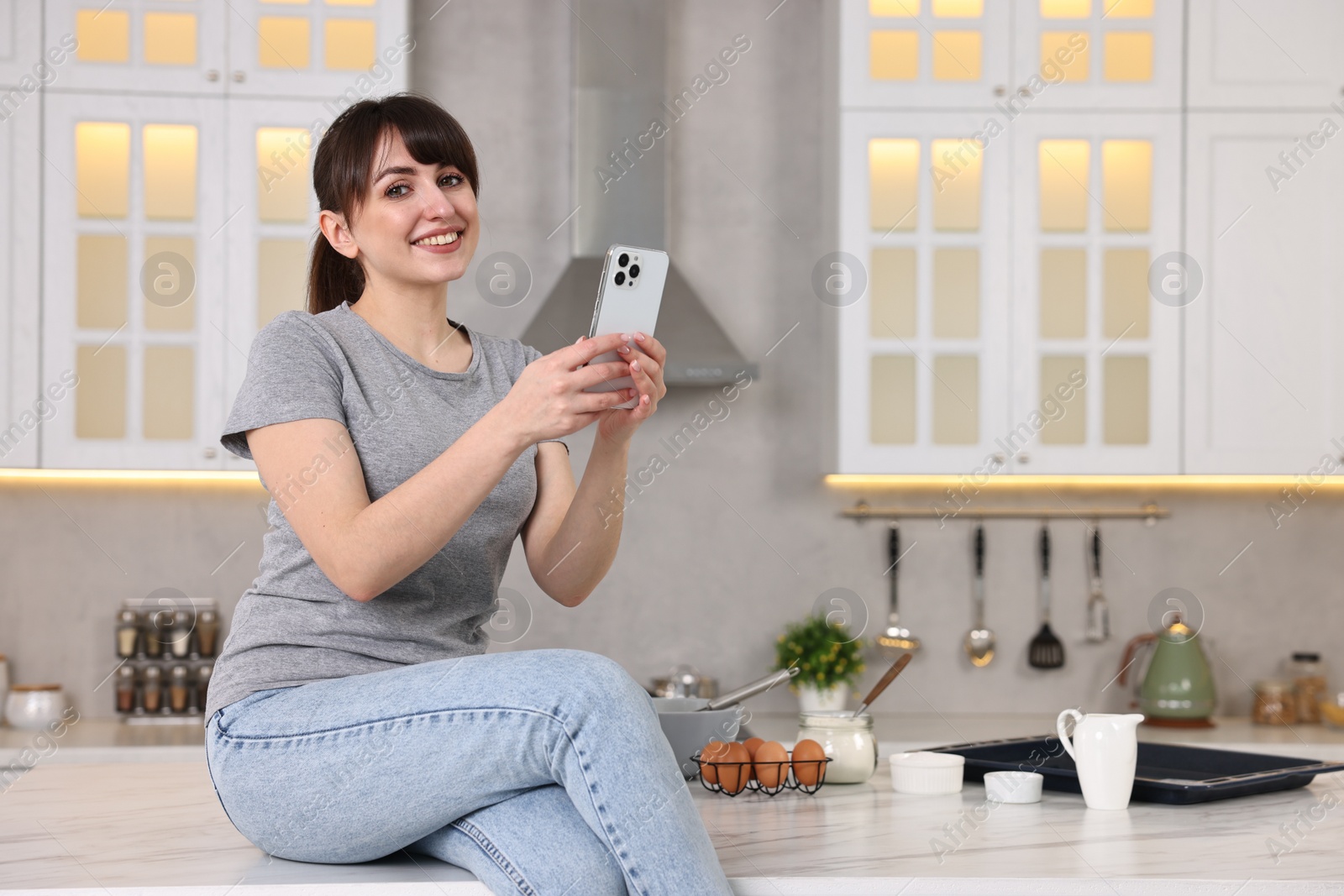 Photo of Happy young housewife using smartphone on white marble table in kitchen