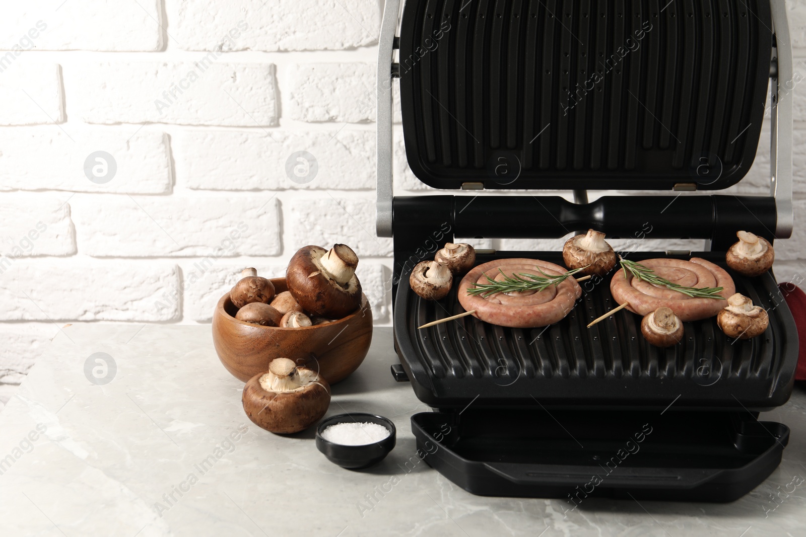 Photo of Electric grill with homemade sausages, rosemary and mushrooms on marble table. Space for text