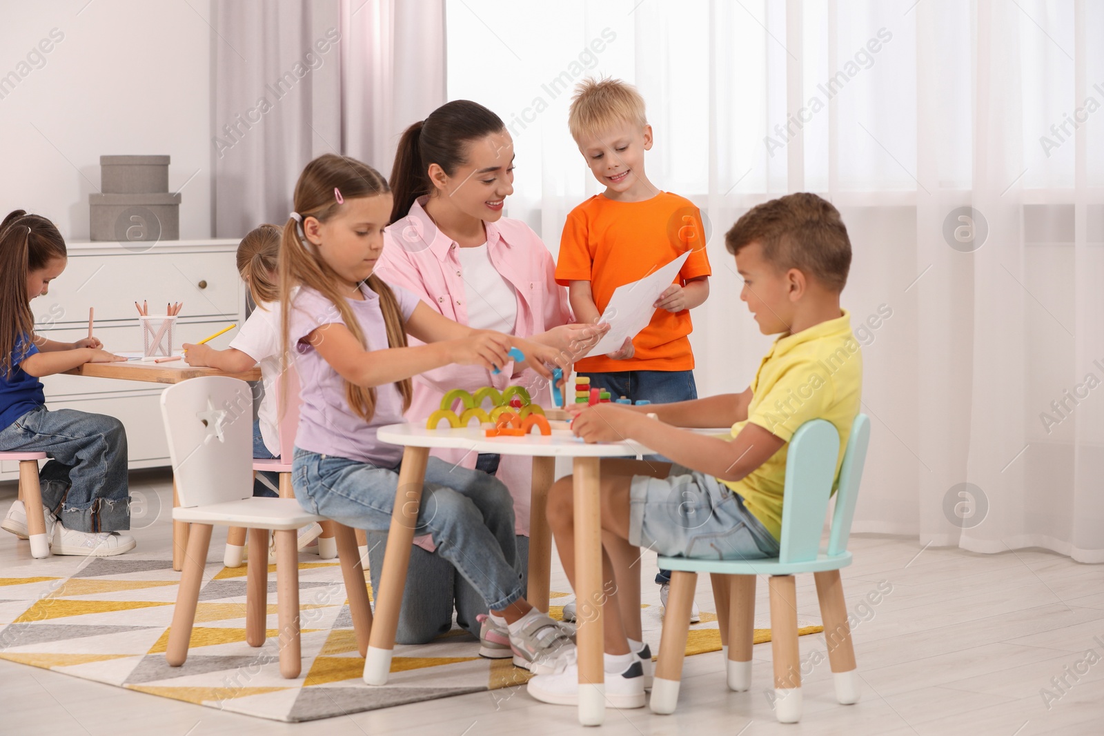 Photo of Nursery teacher and group of cute little children playing at desks in kindergarten. Activities for motor skills development