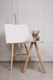 Easel with blank canvas and different art supplies on wooden table near white wall indoors