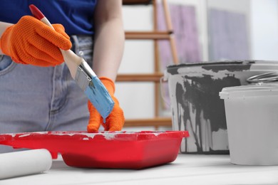 Woman taking light blue paint with brush from tray at white wooden table indoors, closeup