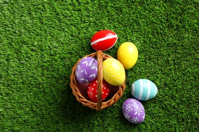 Wicker basket with painted Easter eggs on green grass, top view