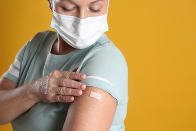 Photo of Mature woman in protective mask showing arm with bandage after vaccination on yellow background