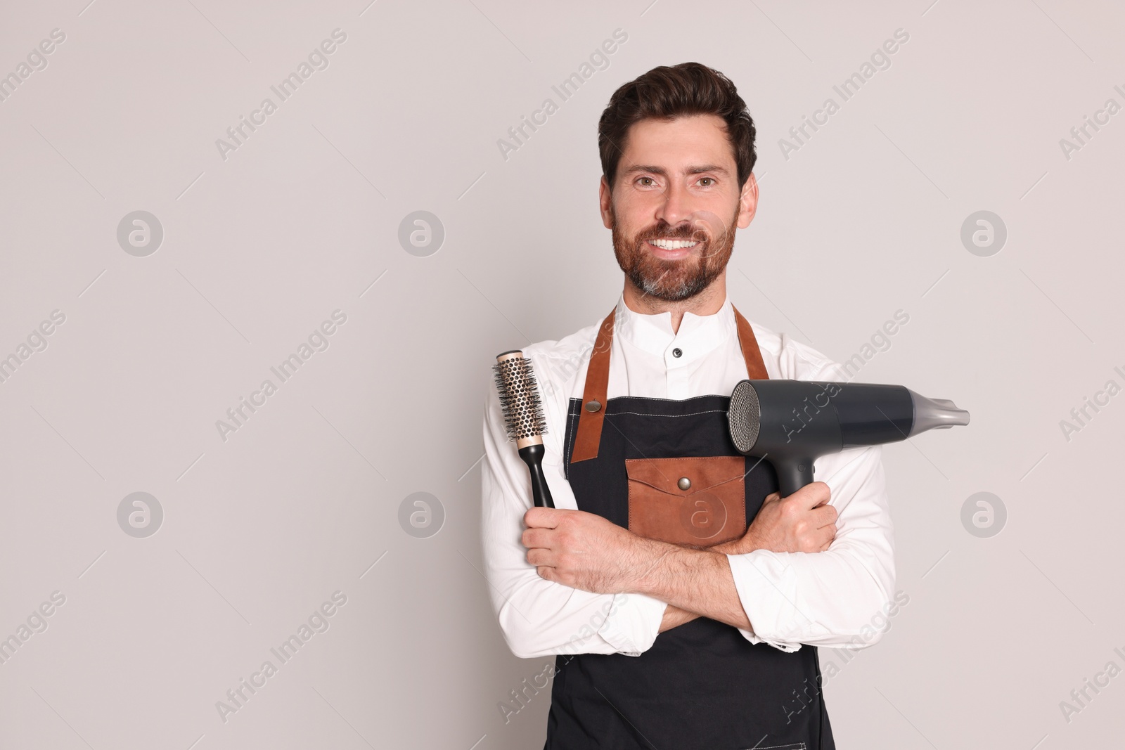 Photo of Smiling hairdresser wearing apron with dryer and brush on light grey background, space for text