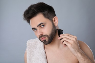 Handsome young man shaving with electric trimmer on grey background