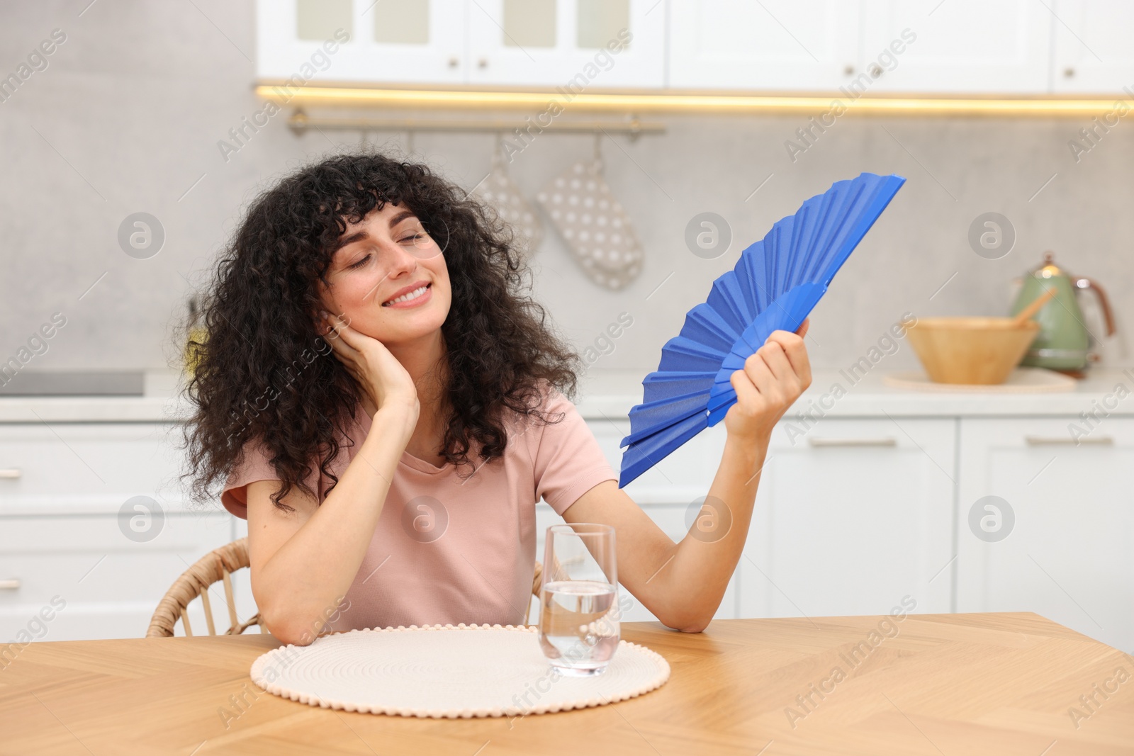 Photo of Young woman waving blue hand fan to cool herself at table in kitchen