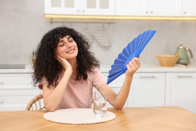 Photo of Young woman waving blue hand fan to cool herself at table in kitchen