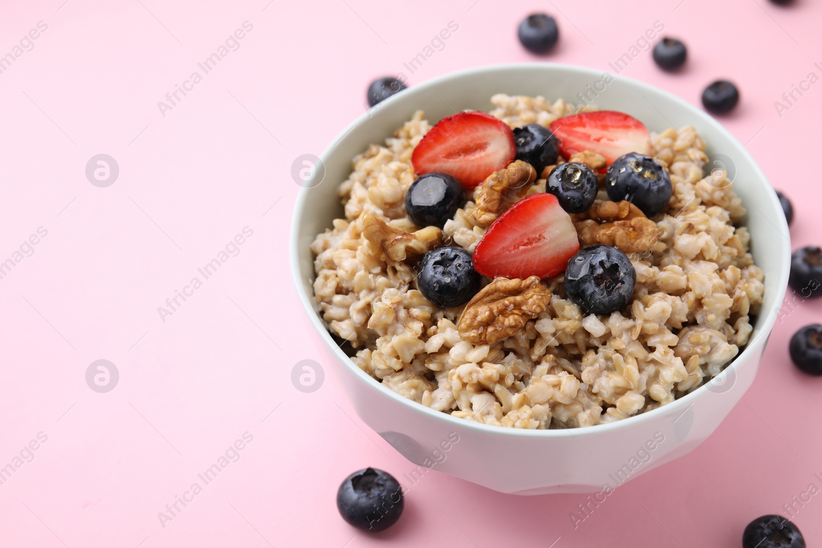 Photo of Tasty oatmeal with strawberries, blueberries and walnuts in bowl surrounded by fresh berries on pink background, space for text