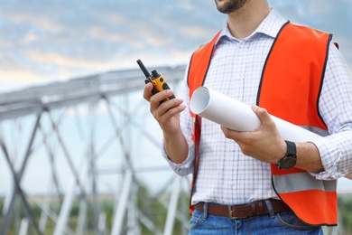 Photo of Professional engineer with plan and walkie talkie near high voltage tower construction outdoors, closeup. Installation of electrical substation