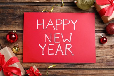 Photo of Happy New Year greeting card. Red paper with phrase and decor on wooden background, flat lay