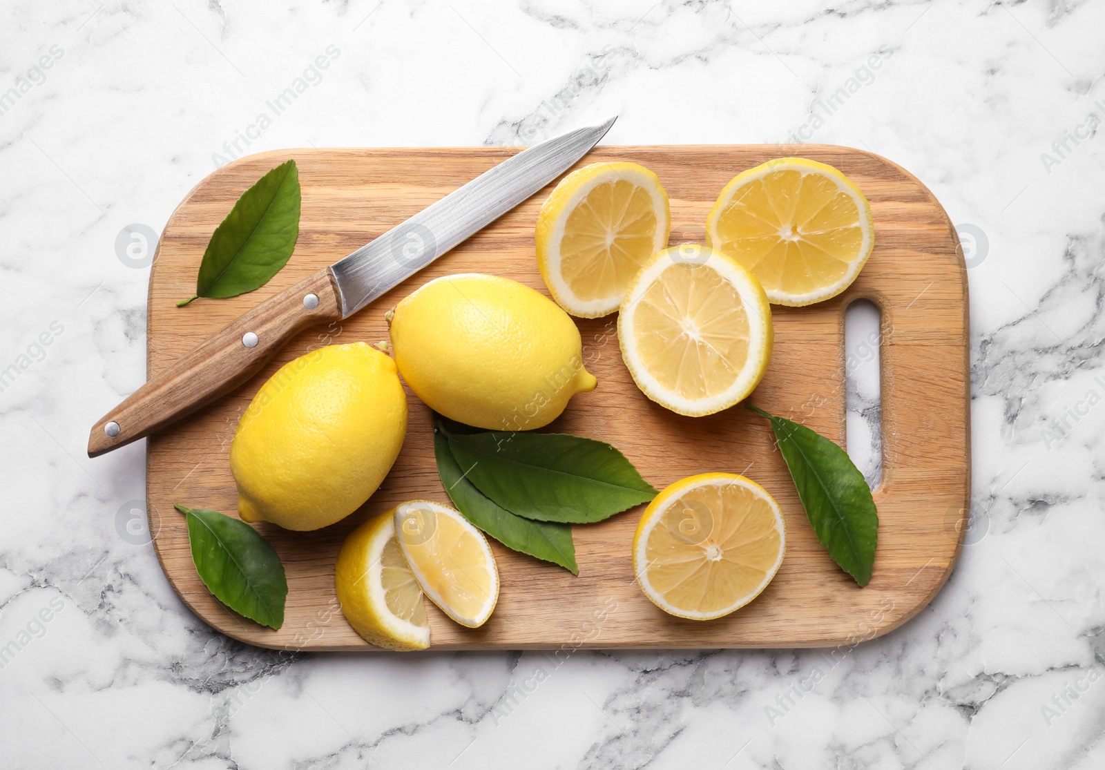 Photo of Many fresh ripe lemons with green leaves and knife on white marble table, top view