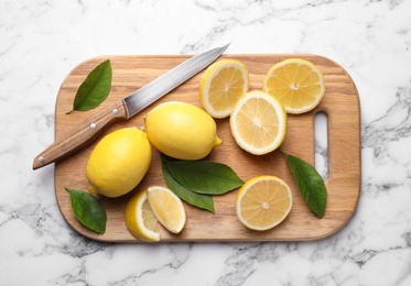 Many fresh ripe lemons with green leaves and knife on white marble table, top view