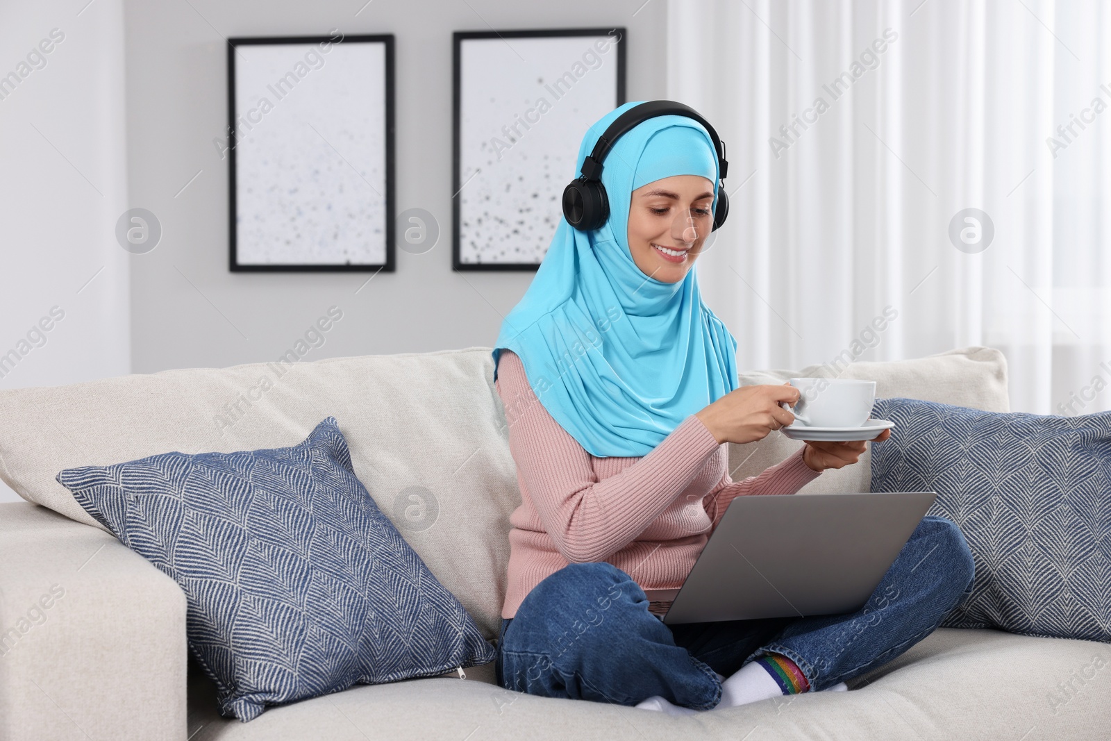 Photo of Muslim woman with cup of drink using laptop at couch in room