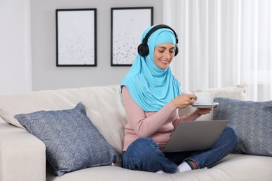 Muslim woman with cup of drink using laptop at couch in room