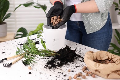Woman in gloves filling flowerpot with drainage at white table indoors, closeup. Repotting houseplants