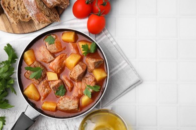 Delicious goulash in saucepan and ingredients on white tiled table, flat lay. Space for text