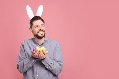 Photo of Happy man in bunny ears headband holding painted Easter eggs on pink background. Space for text