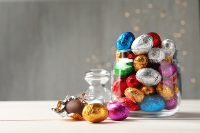 Glass jar with chocolate eggs wrapped in colorful foil on white wooden table. Space for text