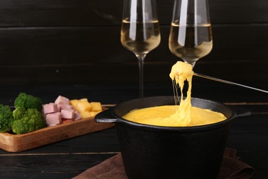 Photo of Dipping piece of ham into fondue pot with melted cheese on black wooden table, closeup